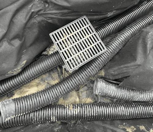 Black perforated drain pipe and a square catch basin grate lying on filter fabric in a trench