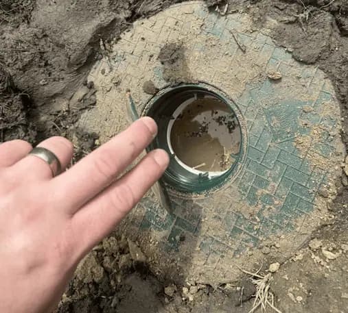 A green pop-up emitter in the soil with its lid lifted, showing the white elbow below