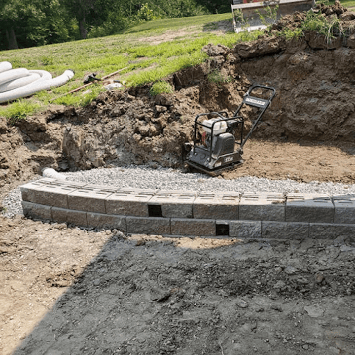 The first course of a block retaining wall on a compacted gravel base, with a plate compactor and drain pipe on site