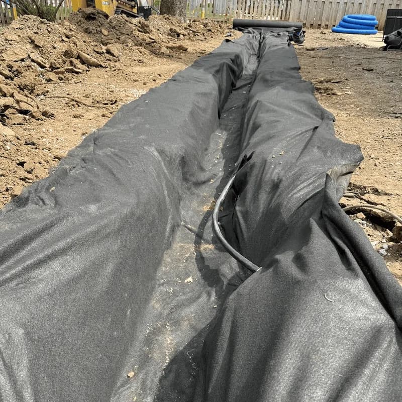 Black filter fabric laid along the full length of an excavated French drain trench