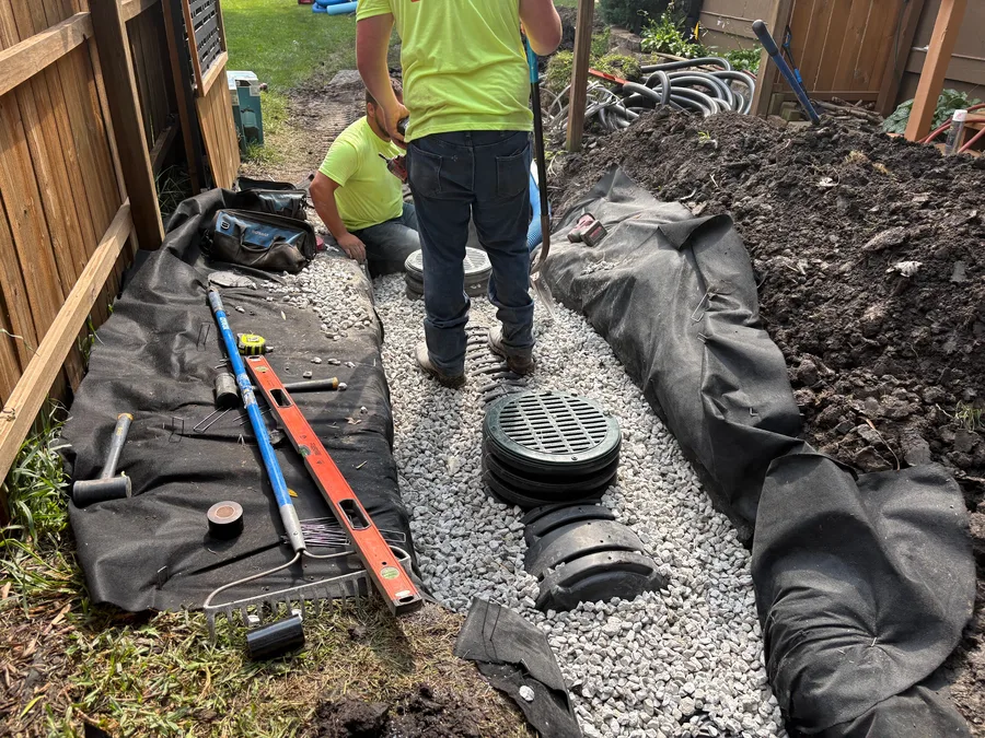 Outdoor sump basin set in washed gravel between fabric-lined trench walls, installer levelling the rim